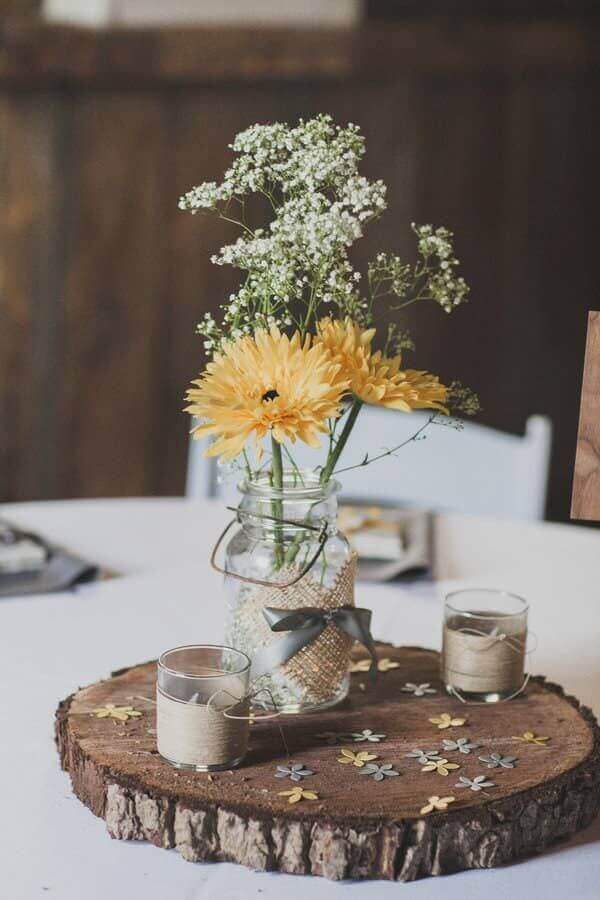 Estilo Country: Centro de mesa rústico con flor amarilla en tarro de vidrio, cinta de lino y base de tronco decorado con mariposas