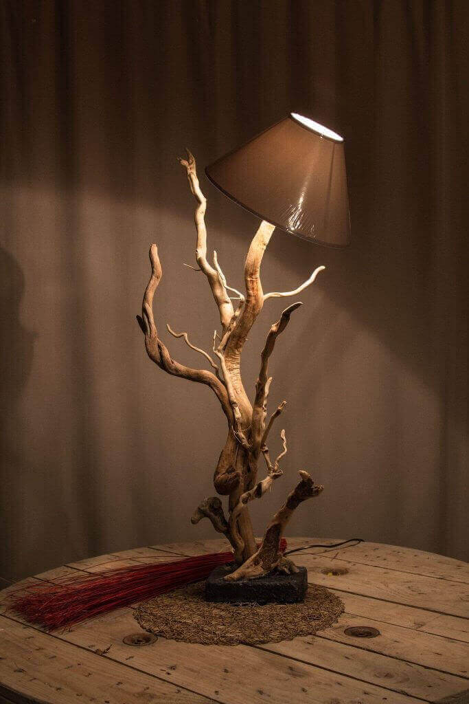 Lámpara con base de ramas retorcidas sobre mesa rústica con objeto decorativo rojo.